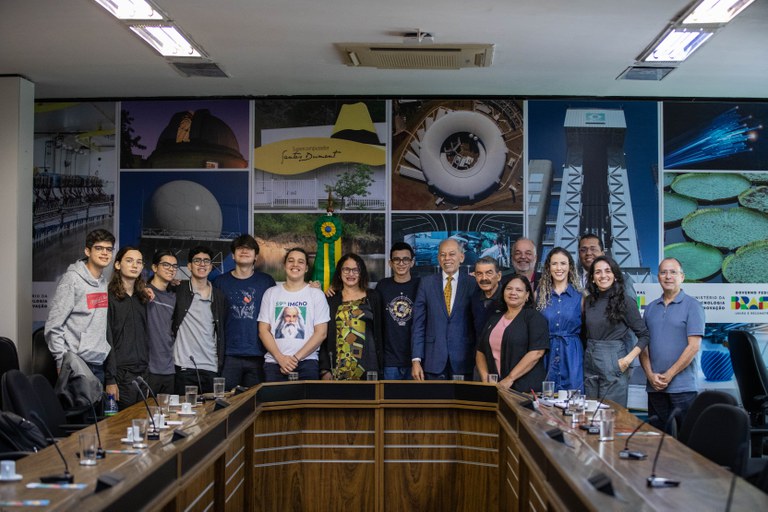 Um grande grupo de estudantes e adultos sorrindo ao redor de uma mesa de conferência.