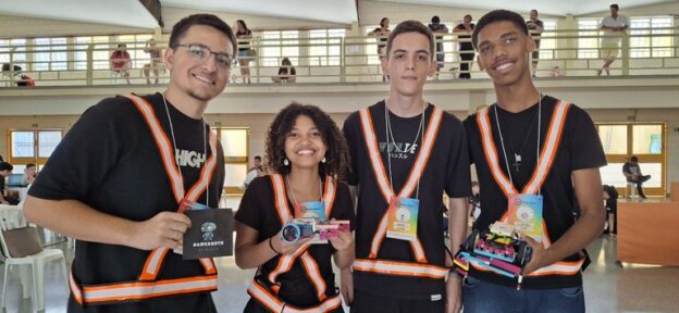 Quatro jovens sorrindo, vestindo coletes laranja e segurando partes de robôs, em um evento indoor.