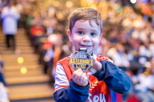 Criança sorridente segurando uma medalha do programa "Caça Asteroides MCTI".